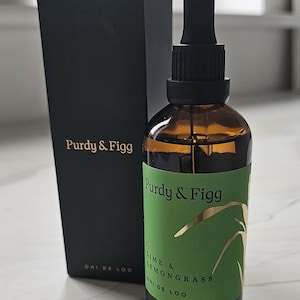以下が含まれることがあります： 黒いスポイトキャップが付いた琥珀色のガラス瓶と、Purdy & Figg Lime & Lemongrass Ohi De Looという文字が書かれた緑色のラベル。その隣には、金色の文字でPurdy & FiggとOhi De Looと印刷された背の高い黒い長方形の箱が置かれています。