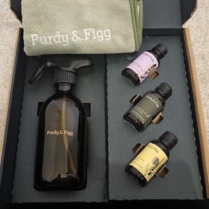 Könnte beinhalten: Ein offenes Box-Set mit Reinigungsprodukten. Es enthält eine dunkelbraune Sprühflasche, ein grünes Mikrofasertuch mit "Purdy & Figg"-Aufdruck und drei kleine Flaschen. Die Box enthält auch Anweisungen.