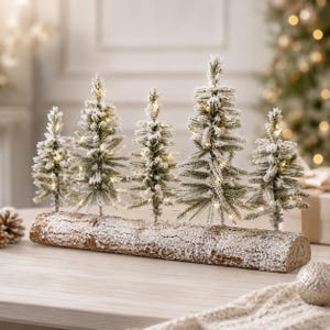 Puede incluir: Un centro de mesa decorativo con cinco árboles de hoja perenne artificiales con un acabado flocado que simula nieve, dispuestos en fila sobre una base de tronco de madera larga y texturizada. Cada árbol tiene luces LED cálidas.