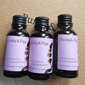 Könnte beinhalten: Drei bernsteinfarbene Glasflaschen mit schwarzen Schraubverschlüssen, jede mit einem hellvioletten Etikett mit dem Text Purdy & Figg, Patchouli & Cedarwood und Counter Clean, in einer Reihe vor einem Kartonhintergrund.