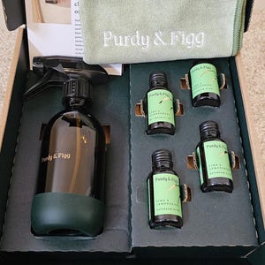 Könnte beinhalten: Eine offene Box mit einer schwarzen Sprühflasche, kleinen grünen Flaschen und einem grünen Tuch. Die Flaschen sind mit "Purdy & Figg" und "Lime & Lemongrass Bathroom Bliss" beschriftet. Das Tuch ist mit "Purdy & Figg" bestickt.