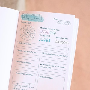 Op de afbeelding: Open journaalpagina met een "Daily Check-In" lay-out. De pagina bevat secties voor het volgen van stemming, slaapbeoordeling, energieniveaus en waterinname. Het heeft ook prompts voor dagelijkse intenties, dankbaarheid en zelfreflectie.