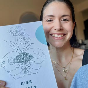Op de afbeelding: Een wit tijdschrift met de woorden "RISE GENTLY" en "A Guided Journal for Your Postpartum Journey" op de omslag. De omslag toont een lijntekening van handen die een plant met bloemen en een lichtblauwe cirkel omarmen.