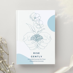 Op de afbeelding: Een wit notitieboek met de woorden "RISE GENTLY" en "A Guided Journal for Your Postpartum Journey" gedrukt op de kaft. De kaft heeft een turquoise lijntekening van handen die een plant met bloemen wiegen. Een turquoise cirkel staat op de achtergrond.