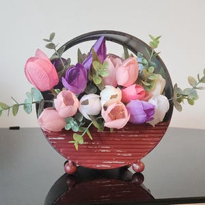 Puede incluir: Una maceta redonda de metal negro con un acabado rojo desgastado contiene un ramo de flores artificiales. Las flores son rosas, moradas y blancas y tienen un aspecto realista.