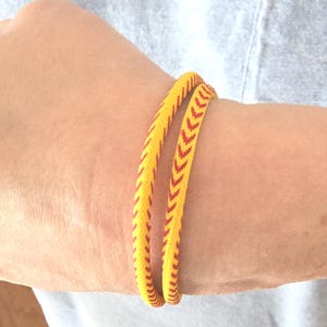 May include: Two yellow bracelets with red chevron patterns are worn on an arm. The bracelets are made of a woven material and have a braided appearance. The bracelets are a bright yellow colour with a repeating red chevron design.