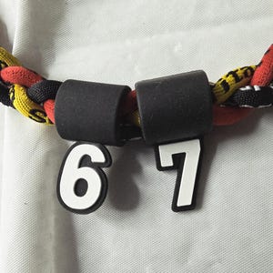 May include: A close-up of a braided necklace with black, red, and yellow strands. Two black cylindrical beads connect the necklace. White numbers 6 and 7 are attached to the necklace.