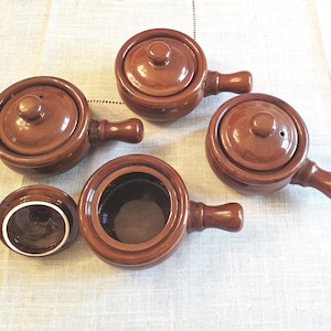 May include: A set of four small, round, brown glazed ceramic pots with matching lids and side handles. Three pots are covered with their lids, while one pot is shown open with its lid placed separately to the side, revealing a dark interior.