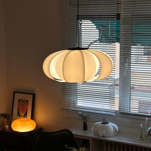 Könnte beinhalten: Eine cremefarbene, oval geformte Pendelleuchte hängt von der Decke. Das Licht hat ein segmentiertes Design mit einem dunklen, kreisförmigen Sockel. Der Raum verfügt außerdem über eine kleine Tischlampe, ein gerahmtes Kunstwerk und ein Fenster mit Jalousien.