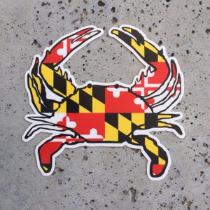 May include: A white crab silhouette with the Maryland state flag design in red, yellow, and black. The crab is facing right with its claws raised.