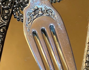 May include: A silver fork with a floral design has been transformed into a pendant necklace. The fork is attached to a silver chain.
