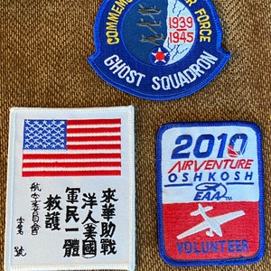 May include: Three embroidered patches. The first is blue with white text that reads "Commemorative Air Force Ghost Squadron" and includes the years "1939-1945". The second is a white patch with a red, white, and blue American flag and text in black. The third is red, white, and blue with the text "2010 Airventure Oshkosh EAA* Volunteer".