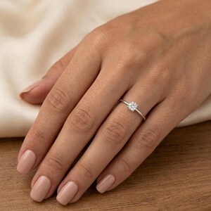 May include: A delicate silver ring featuring a small, round, clear gemstone. The ring is set on a thin band and is displayed on a hand with manicured nails. The background is a soft, neutral color.