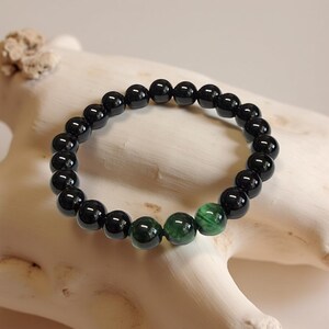 May include: A beaded bracelet featuring black and green gemstones. The bracelet is composed of polished, round beads. The majority of the beads are black, with a few green beads interspersed. The bracelet is displayed on a white, textured surface.