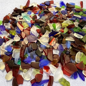 May include: A pile of sea glass in various colors, including red, blue, green, white, and brown. The pieces are irregular in shape and size, with smooth, frosted surfaces. The sea glass is scattered on a light blue surface.
