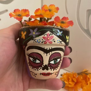 May include: A small terracotta flower pot painted with a Day of the Dead sugar skull face. The pot is decorated with black, white, purple, pink, and green paint. The pot is filled with orange flowers.