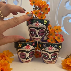 May include: Three small terracotta flower pots painted with colorful Day of the Dead sugar skull faces. Each pot has a black rim with a burlap band and is decorated with colorful flowers. The pots are stacked on top of each other.