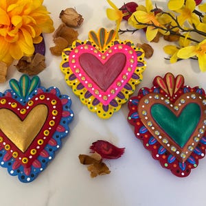May include: Three vibrantly painted heart-shaped ornaments. The ornaments feature gold, pink, and green hearts with decorative borders in red, blue, and yellow. Each heart is adorned with small, white dots. Yellow flowers and dried petals are in the background.