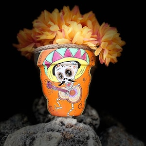 May include: A terracotta flower pot painted with a Day of the Dead skeleton playing a guitar. The skeleton is wearing a sombrero and has a mustache. The pot is decorated with orange, yellow, green, and pink paint. The pot is sitting on a rock with yellow flowers.