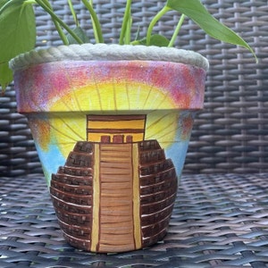 May include: A terracotta flower pot painted with a colorful scene of a Mayan pyramid with a yellow sun shining down. The pot has a rope rim.