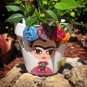 Puede incluir: Maceta de cerámica blanca pintada con un retrato de Frida Kahlo con una corona de flores de colores. La maceta tiene una planta verde que crece fuera de ella y una pequeña pegatina de un mono en la parte delantera.
