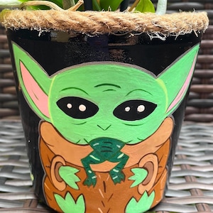 May include: A black painted ceramic flower pot featuring a green character with large eyes and pointed ears. The character is eating a green frog. The pot has a brown rim and is decorated with a rope.
