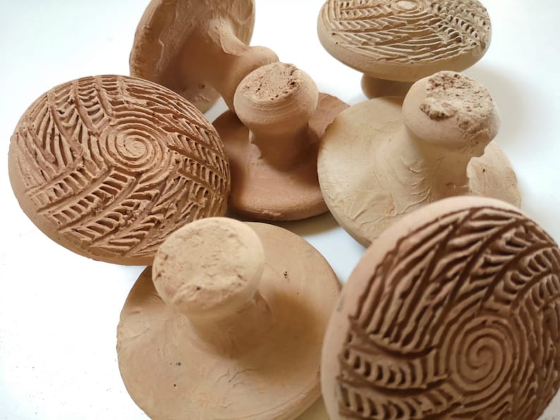 Puede incluir: Seis sellos de arcilla de terracota con diferentes dise&ntilde;os. Los sellos tienen forma de hongo con una parte superior redonda y un tallo corto. Las partes superiores de los sellos tienen patrones intrincados, que incluyen espirales y l&iacute;neas.