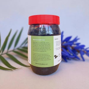 Puede incluir: Un tarro de jab&oacute;n Beldi marroqu&iacute; con tapa roja. El tarro tiene una etiqueta verde y blanca con texto que describe los ingredientes y beneficios del jab&oacute;n. El jab&oacute;n es de color marr&oacute;n oscuro y parece gel. El fondo incluye hojas verdes y flores azules.