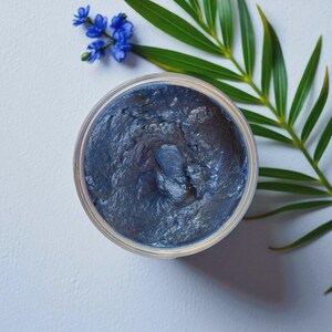Puede incluir: Un tarro abierto de producto cosm&eacute;tico azul oscuro, posiblemente una mascarilla o crema facial, se encuentra sobre una superficie blanca. La superficie texturizada del producto es visible. Flores azules y una hoja de palma verde a&ntilde;aden un toque natural.