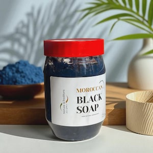 Op de afbeelding: Een pot Marokkaanse zwarte zeep met een rode deksel en een wit etiket. Op het etiket staat "MOROCCAN BLACK SOAP" met het merk "Nature & Traditions". Op de achtergrond staat een kom met blauw poeder.