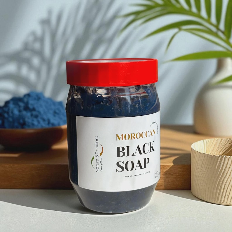 Puede incluir: Un tarro de jab&oacute;n negro marroqu&iacute; con tapa roja y etiqueta blanca. La etiqueta dice "MOROCCAN BLACK SOAP" con la marca "Nature & Traditions". Un cuenco de polvo azul en el fondo.