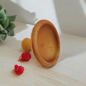 Puede incluir: Un cuenco de madera artesanal con un borde ancho y un acabado natural. El cuenco es de color marrón claro con un borde dorado. Dos pétalos de flores rojas y una fruta amarilla están junto al cuenco.