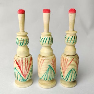 May include: Three decorative wooden objects with a light beige finish. Each has a red top, a long neck, and a base. The bodies are decorated with red and green painted lines. The objects are displayed on a white surface.