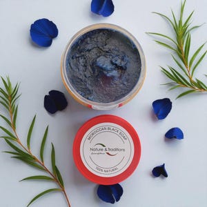 Puede incluir: Vista a&eacute;rea de un tarro de jab&oacute;n negro marroqu&iacute; con la tapa quitada, que revela el jab&oacute;n azul oscuro. La tapa roja est&aacute; al lado del tarro, con el texto "MOROCCAN BLACK SOAP" y "Nature & Traditions" impreso. P&eacute;talos azules y hojas verdes rodean el tarro.