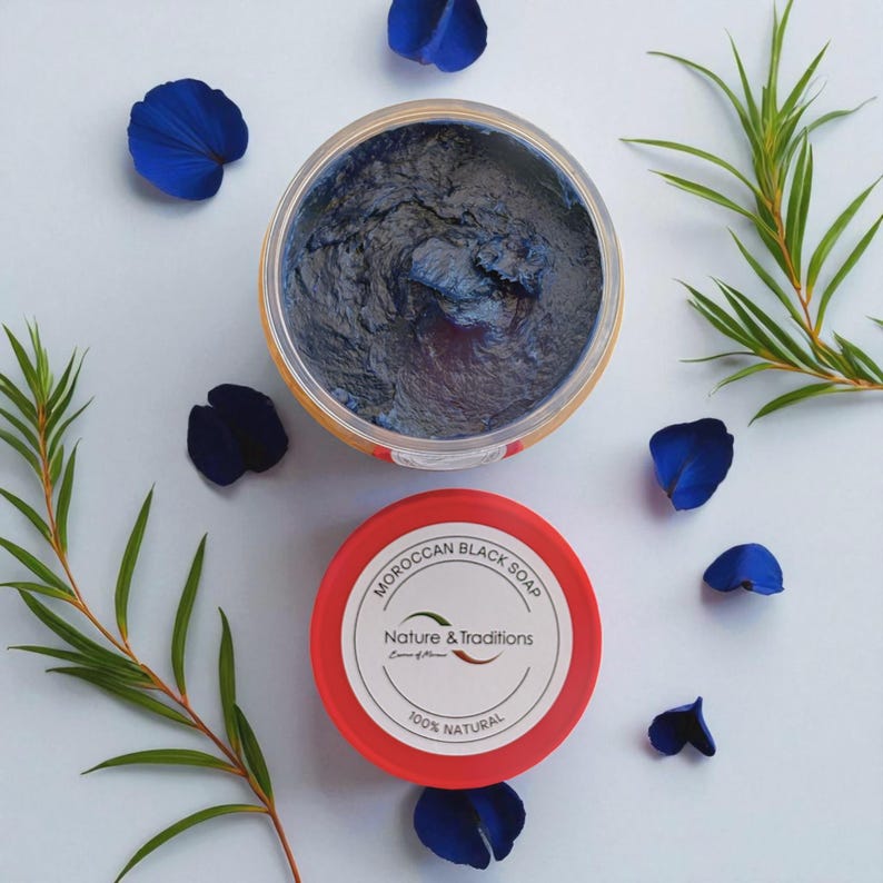 Puede incluir: Vista a&eacute;rea de un tarro de jab&oacute;n negro marroqu&iacute; con la tapa quitada, que revela el jab&oacute;n azul oscuro. La tapa roja est&aacute; al lado del tarro, con el texto "MOROCCAN BLACK SOAP" y "Nature & Traditions" impreso. P&eacute;talos azules y hojas verdes rodean el tarro.