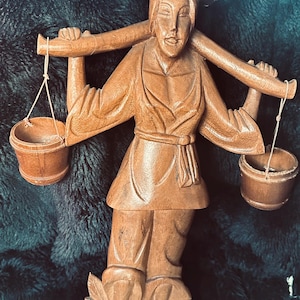 May include: A wooden figurine depicting a person carrying two buckets suspended from a pole. The figure wears a conical hat and traditional clothing. The carving is detailed, with visible wood grain and a natural finish.
