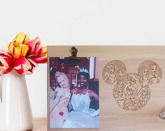 May include: A wooden photo frame with a clip to hold a photo. The frame has a Mickey Mouse silhouette carved into the wood, with the silhouette made up of Disney princesses. The photo in the frame shows two girls dressed as princesses.