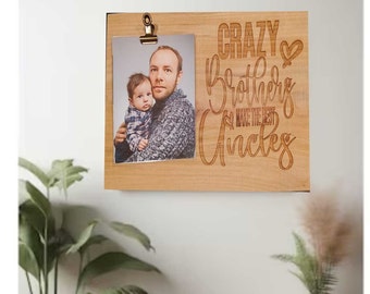 May include: A wooden photo frame with a clip at the top. The frame has a photo of two men, one holding the other. The text "Crazy Brothers Make The Best Uncles" is engraved on the frame.