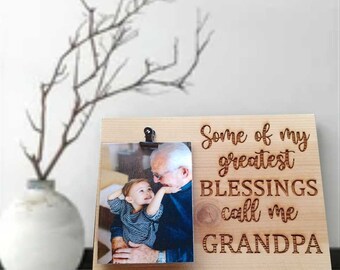 May include: A wooden photo frame with a clip to hold a photo. The frame has the text "Some of my greatest BLESSINGS call me GRANDPA" engraved on it. A photo of an older man and a young child is in the frame.