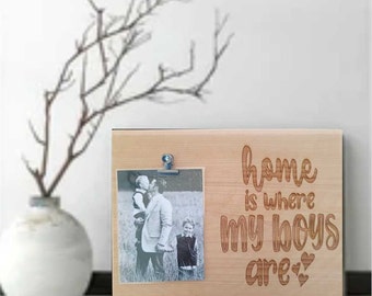 May include: A wooden photo frame with the text "home is where my boys are" engraved on it. The frame has a clip to hold a photo, and a black and white photo of a father and his two sons is displayed in the frame.