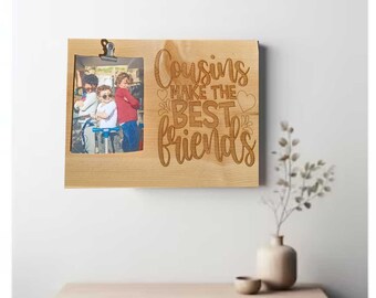 May include: Wooden photo frame with a cutout for a picture and the text "Cousins make the best friends" engraved on the front. The frame is light brown and has a clip at the top.