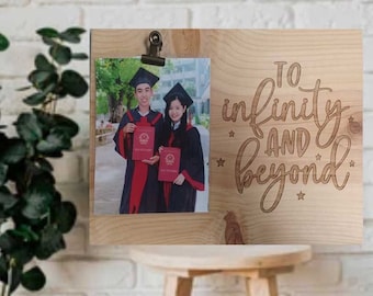 May include: A wooden photo frame with a clip to hold a photo. The frame has the text "To infinity and beyond" engraved on it with stars. The photo shows two people in graduation robes.