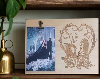 May include: A wooden picture frame with a cutout design of Jack Skellington and Sally from The Nightmare Before Christmas. The frame has a clip to hold a photo, and the photo shows a couple in a wedding setting.