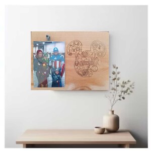 May include: A wooden picture frame with a Mickey Mouse silhouette design engraved on the surface. The frame has a photo of a family dressed as superheroes, including Captain America, Iron Man, and Thor. The frame is hanging on a white wall.