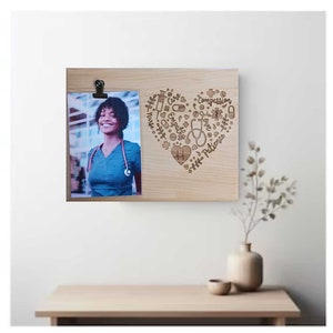 May include: A wooden photo frame with a heart-shaped design engraved with words like "Care", "Compassion", "Love", and "Patience". The frame has a photo of a woman wearing a blue scrubs top and a stethoscope.