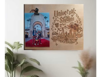 May include: A wooden photo frame with a clip to hold a photo. The frame is engraved with the words "Universal Studios" and a design featuring characters from the theme park. A photo of a family standing in front of the Universal Studios Hollywood entrance is displayed in the frame.