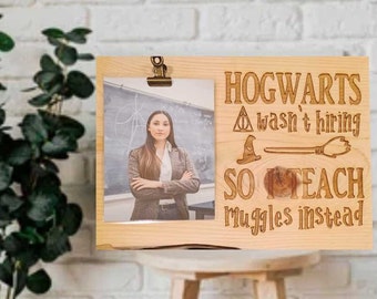 May include: A light-colored wooden picture frame with a photo of a woman in a blazer. The frame has the text "HOGWARTS wasn't hiring SO TEACH Muggles instead" with etched symbols.