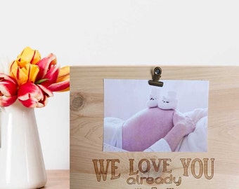 May include: A wooden picture frame with a photo of a pregnant woman's belly with baby booties on top. The frame has the text "WE LOVE YOU already" engraved on it.