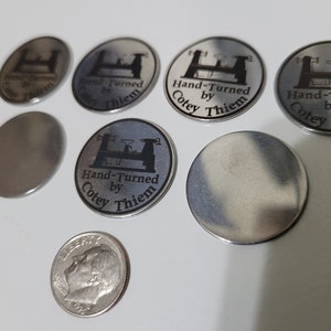 May include: A collection of silver metal discs with a black engraved design. The design features a lathe and the text "Hand-Turned by Cotey Thiem".