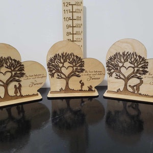 Puede incluir: Tres placas de madera en forma de corazón con un árbol y dos figuras de pie debajo. Cada placa tiene una inscripción diferente: "El amor entre una madre y una hija es para siempre", "El amor entre una madre y un hijo es para siempre" y "El amor entre una madre y un hijo es para siempre".